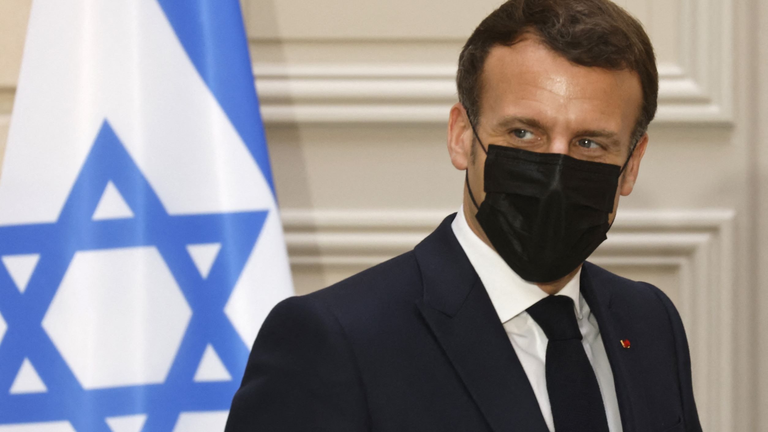 الصورة تظهر رجلًا يرتدي بدلة رسمية وقناعًا أسود. خلفه يوجد علم يحمل نجمة داود. يبدو الرجل في وضعية جانبية، حيث يلتفت نحو الجانب. تعكس الإضاءة في الصورة أجواء رسمية. لا يمكنني تحديد من هو هذا الشخص.