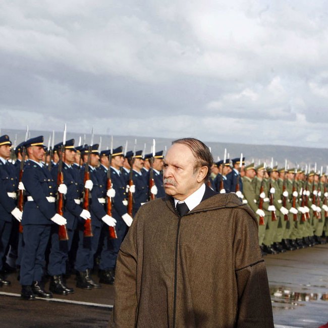 L'image montre un homme en manteau se tenant devant un alignement de soldats en uniforme. Les soldats, majestueux et bien rangés, saluent. L'arrière-plan est nuageux, créant une atmosphère cérémonielle. Le sol semble humide, indiquant peut-être qu'il a plu récemment. L'homme au premier plan a une expression pensive, ajoutant un ton sérieux à la scène.