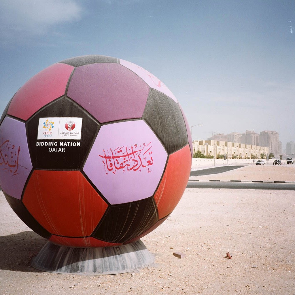 L'image montre un énorme ballon de football décoré, affichant des inscriptions colorées en arabe et d'autres éléments graphiques. Ce ballon est installé sur un terrain, probablement dans un contexte urbain, avec des bâtiments en arrière-plan. Le ciel est clair, et l'environnement semble être une zone en développement ou un lieu préparé pour un événement, suggérant une connexion avec le football ou un événement sportif marquant, tel qu'une compétition internationale.