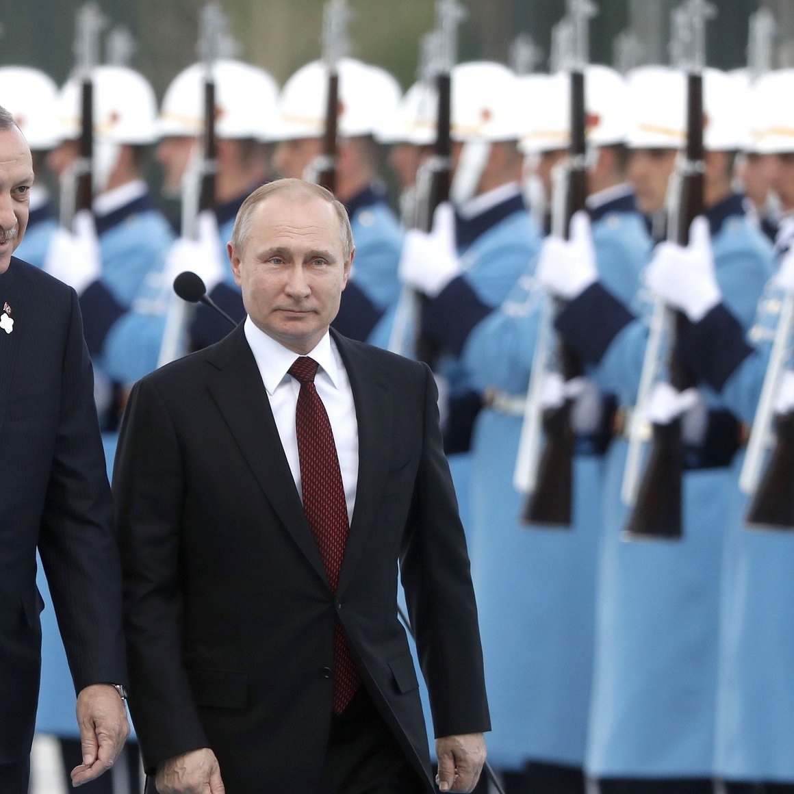 الصورة تظهر شخصين يسيران معًا، بينما خلفهما يظهر مجموعة من الحراس يرتدون زيًا موحدًا، ويقفون في صفوف منتظمة. الشخص الأول يرتدي بدلة داكنة مع ربطة عنق حمراء، بينما الثاني يرتدي بدلة أيضًا ولكن بلون مختلف. يبدو أن المشهد يتمثل في مراسم رسمية أو استقبال، حيث تم ترتيب الحراس بشكل أنيق. الجو العام بالصورة يوحي بالتعاون أو اللقاء بين الشخصيتين.