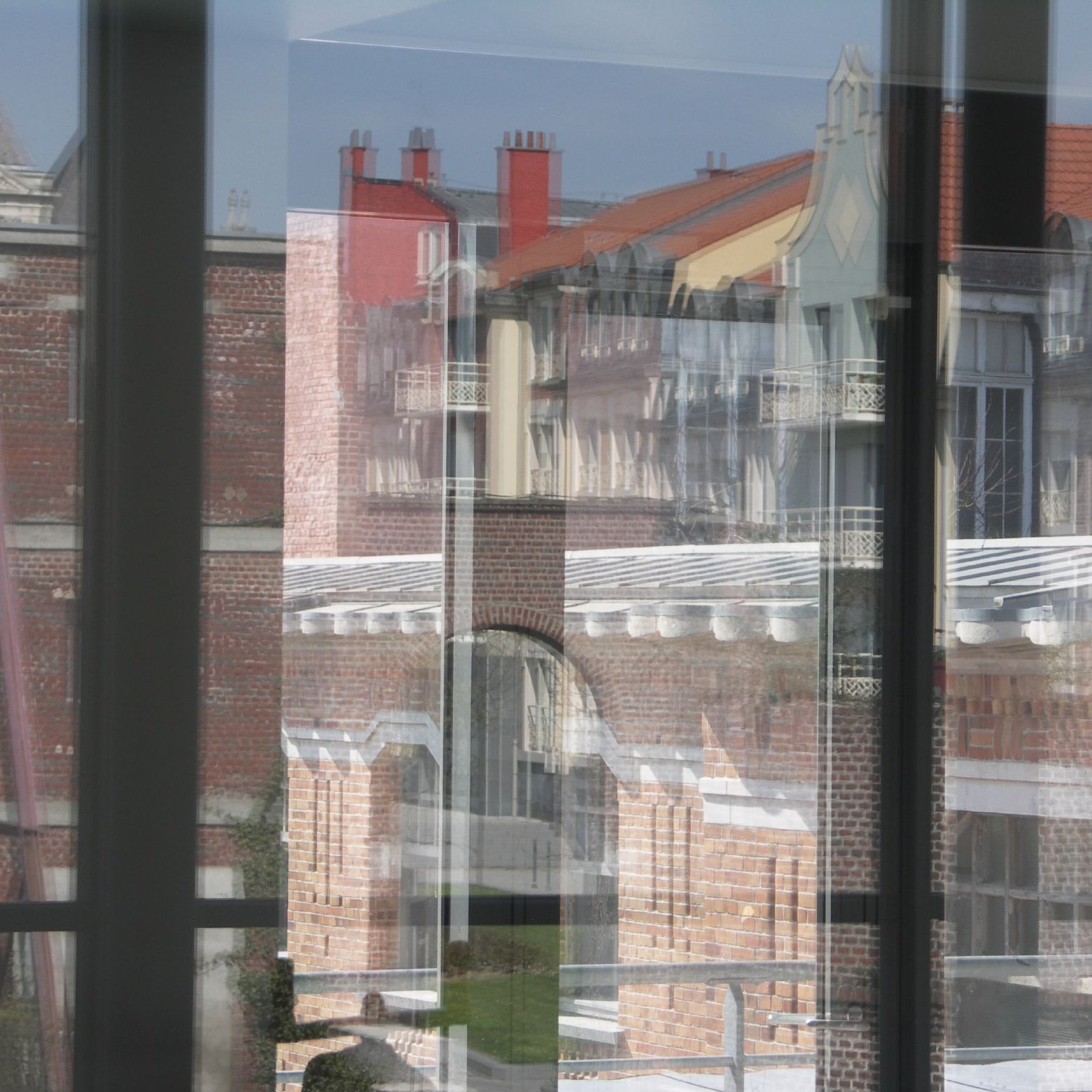 این تصویر نمایی از ساختمان‌ها را از طریق شیشه‌های یک ساختمان دیگر نشان می‌دهد. شیشه‌ها باعث ایجاد انعکاس‌هایی از بیرون می‌شوند که موجب می‌شود جزئیات معماری و رنگ‌های دیوارها و بام‌ها به خوبی نمایان شود. در پس‌زمینه، سقف‌های شیب‌دار و ساختمان‌های مختلف با طراحی‌های متفاوت دیده می‌شود، و نور آفتاب به تصویر جلوه‌ای جذاب می‌بخشد. فضای کلی تصویر حالت مدرنی دارد در حالی که عناصر تاریخی نیز در آن قابل مشاهده است.