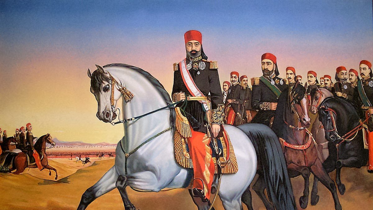 L'image représente un groupe de cavaliers en tenue traditionnelle, menés par un homme à cheval en avant-plan. Cet homme, qui semble être une figure d'autorité, est vêtu d'un uniforme orné et porte un chapeau distinctif. Il est monté sur un cheval gris, tandis qu'un groupe d'hommes sur des chevaux bruns et noirs le suit derrière lui. Le paysage est désertique, avec des montagnes en arrière-plan et un ciel aux teintes chaudes, suggérant le lever ou le coucher du soleil. Cette scène dégage une impression de mouvement et de puissance.