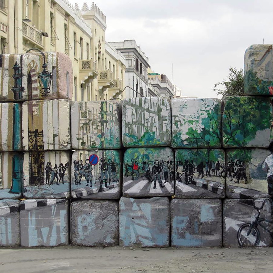 L'image montre un mur de blocs de béton, sur lequel sont peints des paysages urbains. La peinture représente une rue avec des arbres, des piétons et des cyclistes. On peut voir des silhouettes de personnes et des éléments de la ville, comme des lampadaires. Ce contraste entre le mur brut et l'art coloré apporte une touche dynamique à l'environnement urbain. Le ciel est nuageux, ajoutant une atmosphère un peu dramatique à la scène.