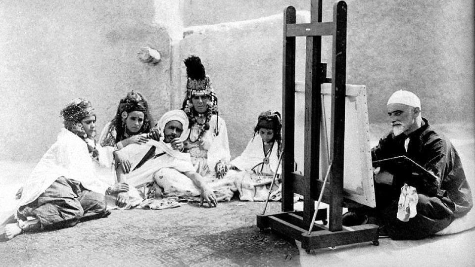 L'image montre un groupe de personnes assises autour d'un homme qui peint sur un chevalet. Les membres du groupe portent des vêtements traditionnels, certains avec des bijoux et des motifs distinctifs. L'homme qui peint semble concentré sur son travail, tandis que les autres l'observent attentivement. L'environnement paraît simple, avec des murs en argile en arrière-plan, ce qui donne un aperçu d'une ambiance culturelle et artistique.