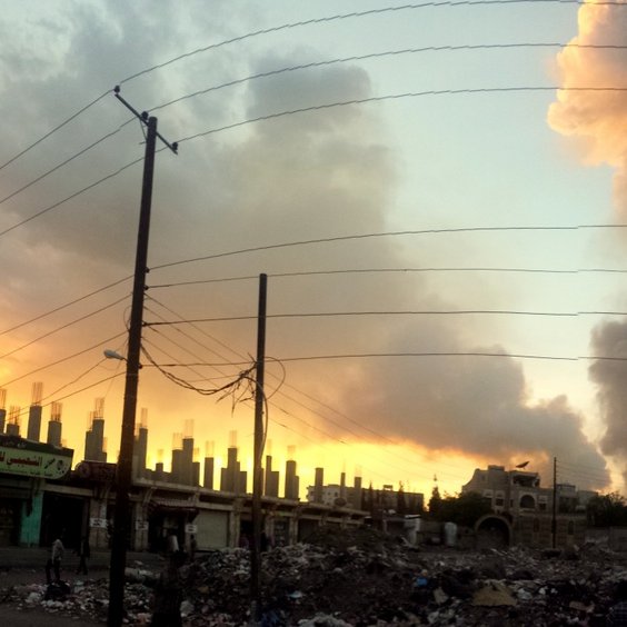 L'image montre un paysage urbain, probablement dans une zone affectée par des conflits. On peut voir des colonnes de fumée s'élevant dans le ciel, indiquant une explosion ou un incendie. À l'arrière-plan, le soleil se couche, créant un contraste entre les couleurs chaudes du ciel et les fumées sombres. Au premier plan, il y a des débris et des déchets éparpillés, et des bâtiments aux murs usés, ornés d'affiches et de signes en arabe. Les fils électriques traversent la scène, renforçant l'impression d'un environnement urbain chaotique.