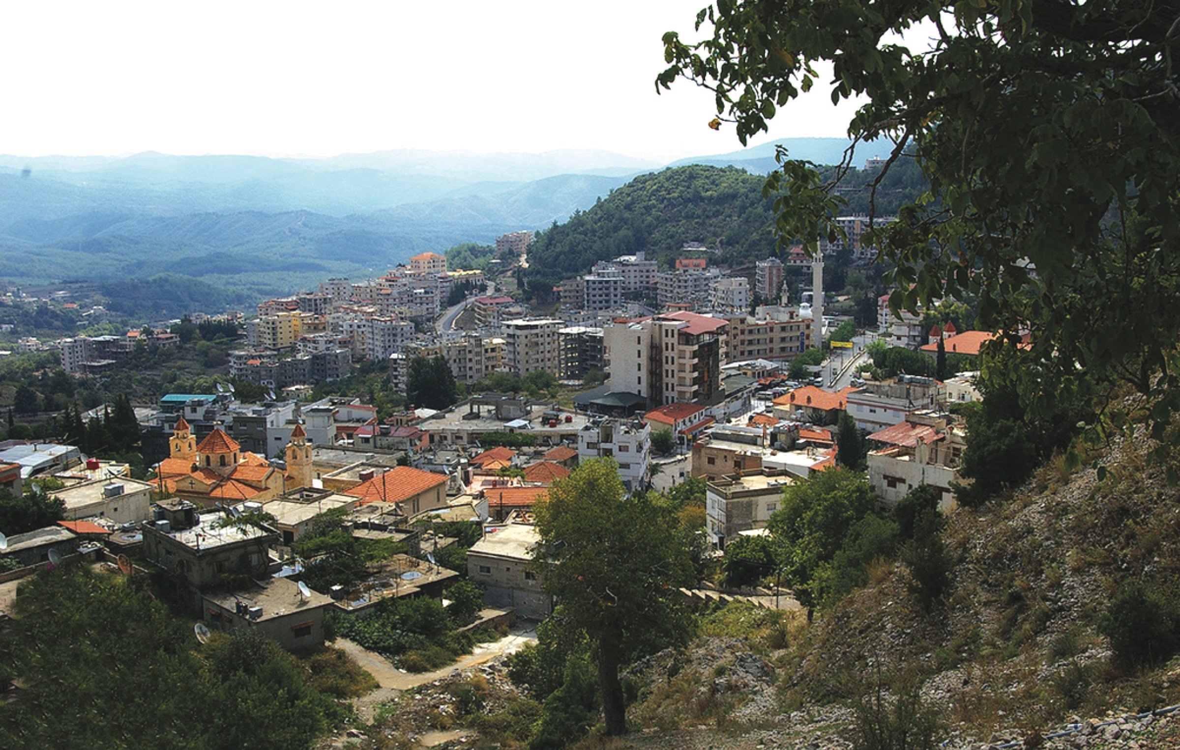 The image shows a panoramic view of a mountainous landscape with a town nestled in the valleys. The foreground features a mix of low-rise buildings, some with red rooftops, and greenery, including trees and hills. In the background, a larger urban area can be seen with taller buildings, likely indicating a more developed part of the town. The sky appears slightly overcast, with mountains extending into the distance, adding depth to the scene. The overall atmosphere suggests a blend of natural beauty and urban development.