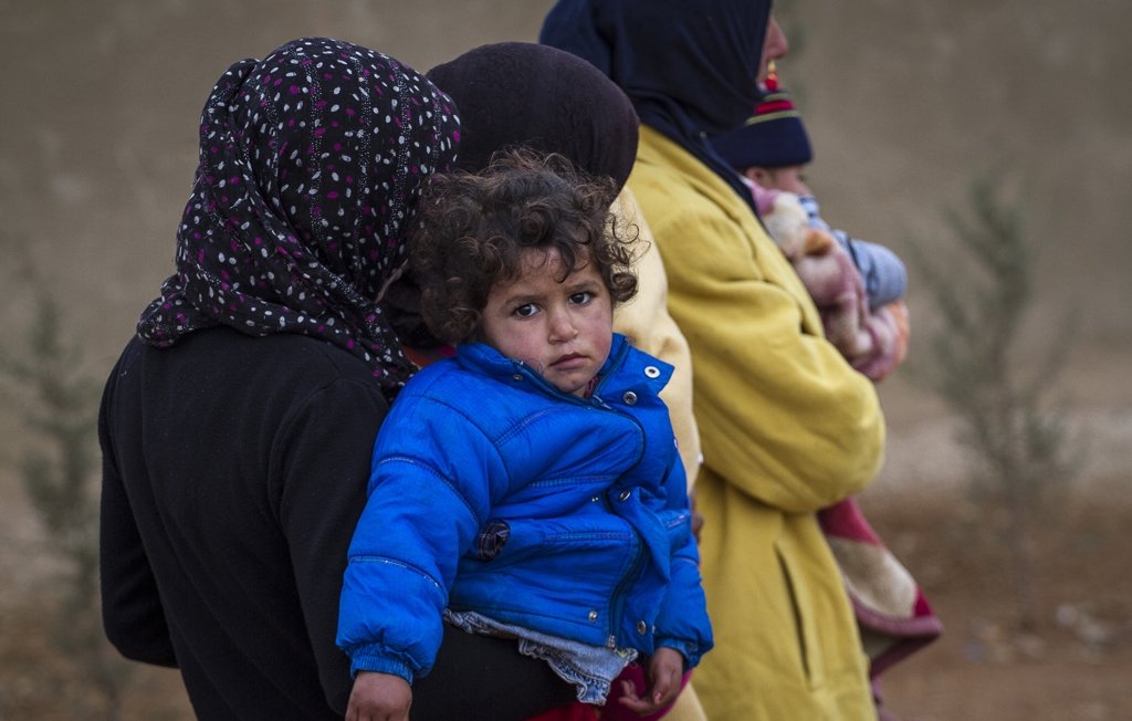 L'image montre plusieurs personnes, dont une femme tenant un enfant dans ses bras. L'enfant porte une veste bleue, et son expression semble triste ou préoccupée. Les femmes qui les entourent sont habillées de manière chaleureuse et portent des foulards. L'arrière-plan est flou, avec des éléments naturels, ce qui suggère un environnement difficile. L'ensemble de la scène évoque une atmosphère de vulnérabilité et de défi.
