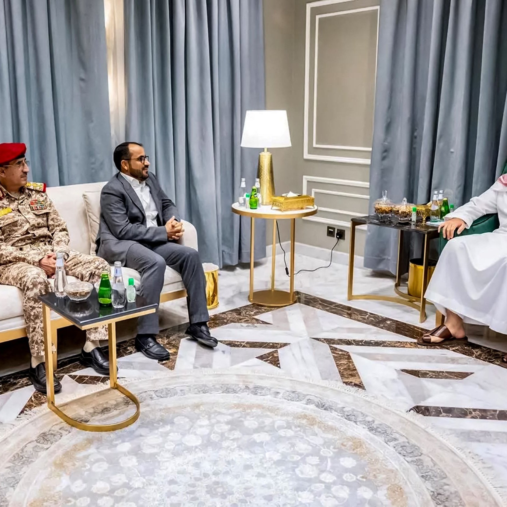 L'image montre une rencontre entre plusieurs hommes assis dans une pièce bien décorée. Deux hommes portent des uniformes militaires, avec des chapeaux rouges, tandis qu'un troisième homme est habillé en costume. Le quatrième homme, à droite, porte une robe traditionnelle. Ils sont assis autour de tables basses où se trouvent des bouteilles d'eau et des verres. Le décor comprend des rideaux gris et un sol en marbre. L'atmosphère semble être celle d'une discussion formelle.