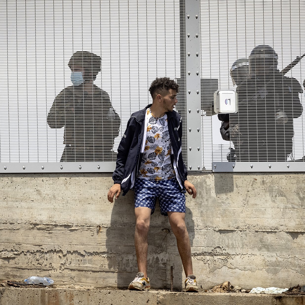 L'image montre un jeune homme debout contre un mur, habillé d'un haut sombre et d'un short à motifs. Il semble être en train de faire face à une situation tendue. Derrière lui, à travers une clôture en métal, on aperçoit des silhouettes de personnes en uniforme, apparemment des agents de la sécurité, qui semblent se préparer à intervenir. L'atmosphère est intense et suggère un contexte de conflit ou de protestation. Les ombres des agents renforcent le sentiment de pression ou de confrontation.