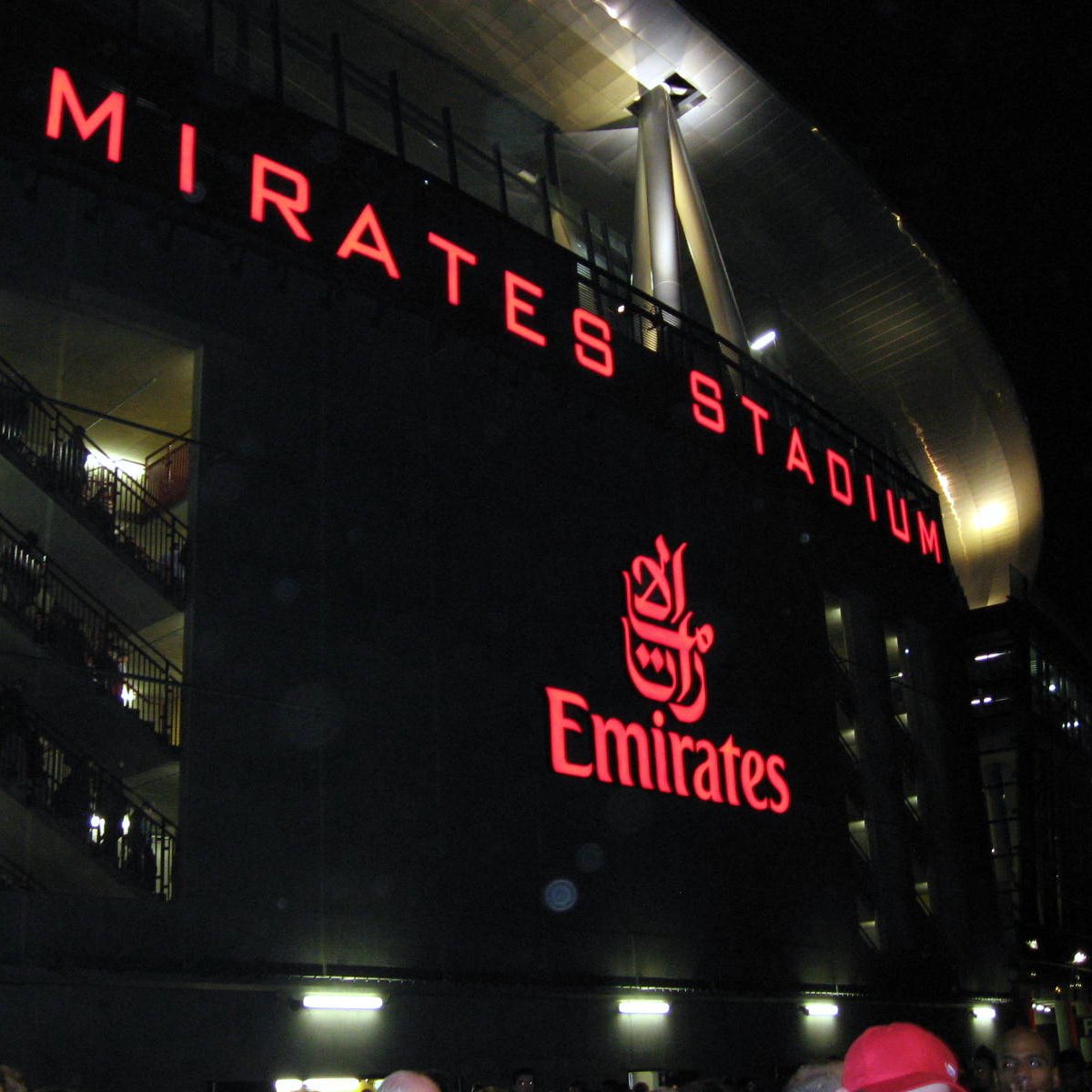 L'image montre l'extérieur de l'Emirates Stadium, qui est la maison du club de football Arsenal, à Londres. Le bâtiment est illuminé, avec le nom "EMIRATES STADIUM" en lettres rouges lumineuses bien visibles. On peut également voir le logo d'Emirates, une compagnie aérienne, mis en évidence. L'ambiance semble animée, suggérant qu'il y a probablement un événement ou un match en cours, avec des personnes rassemblées autour de la façade du stade.