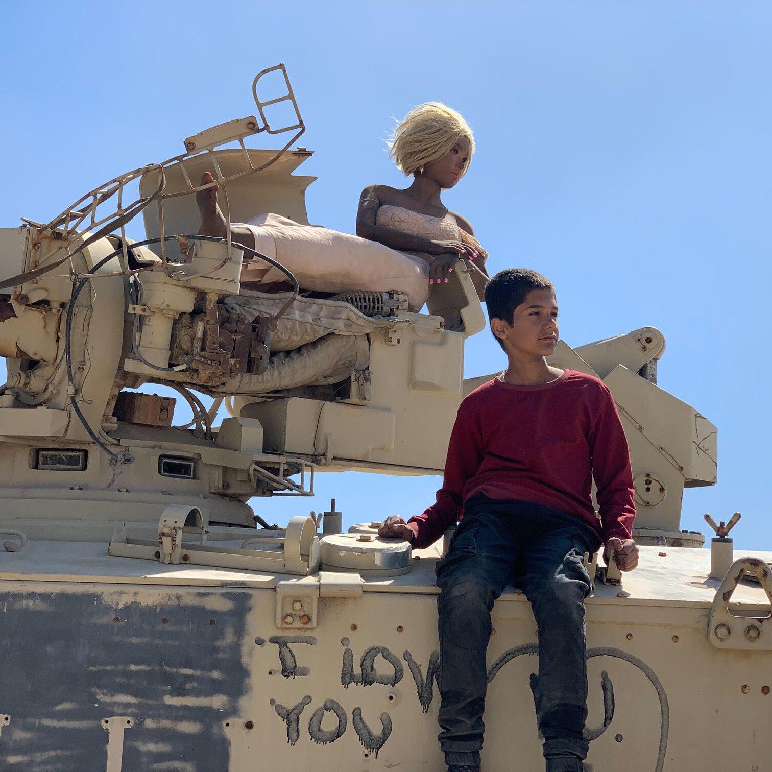 L'image montre un garçon assis sur un véhicule militaire. À côté de lui, il y a une poupée aux cheveux blonds, perchée sur le véhicule. Le fond est clair, avec un ciel bleu. Sur le véhicule, on peut voir des inscriptions, dont "I LOVE YOU" en lettres. L'ensemble de la scène dégage une atmosphere contrastée entre l'innocence de l'enfant et le décor militaire.