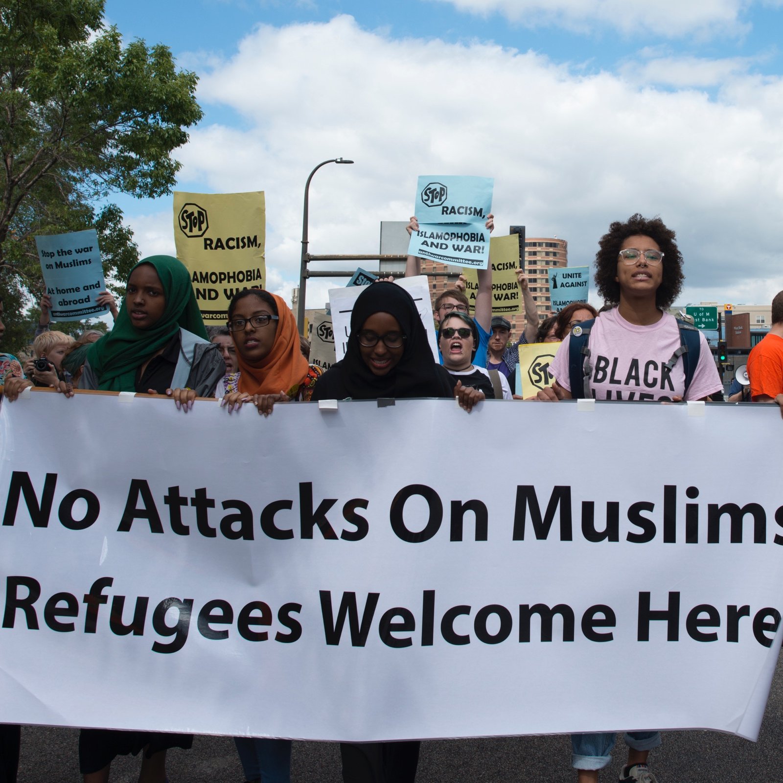 L'image montre un groupe de manifestants en train de marcher dans une rue. Ils portent des pancartes avec des messages tels que "No Attacks On Muslims" et "Refugees Welcome Here". Les participants, majoritairement de culture musulmane, affichent des expressions déterminées. Le ciel est partiellement nuageux, et on peut voir des bâtiments en arrière-plan. L'atmosphère semble engager et militante, soulignant des thèmes de solidarité et de droits humains.
