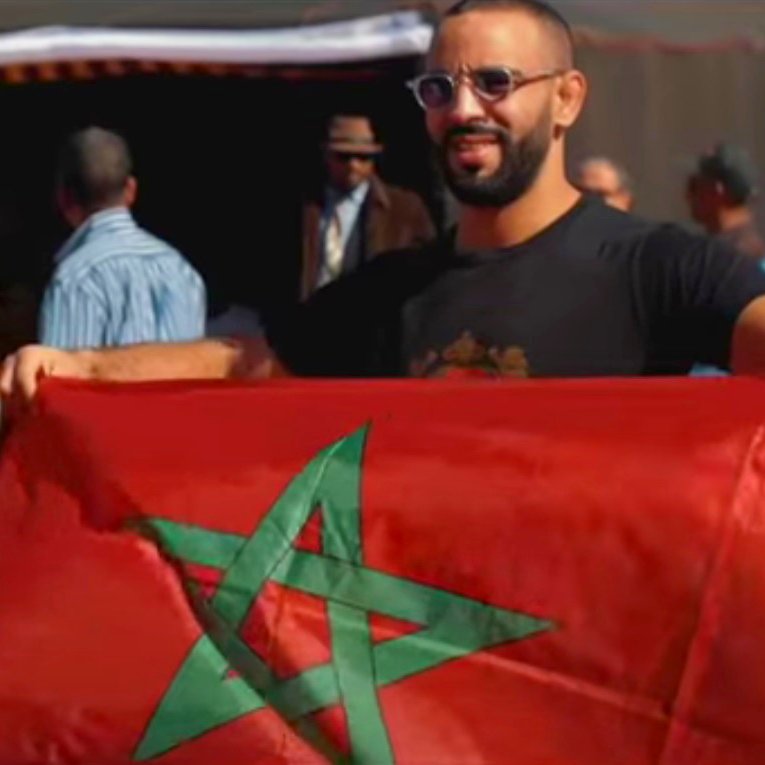 L'image montre un groupe de personnes, dont un homme tenant un drapeau marocain. Il sourit et affiche fièrement le drapeau rouge avec une étoile à cinq branches verte. En arrière-plan, on aperçoit d'autres personnes et des tentes, ce qui laisse supposer une rencontre ou un événement en plein air. L'atmosphère semble festive et conviviale, évoquant un rassemblement culturel ou patriotique.