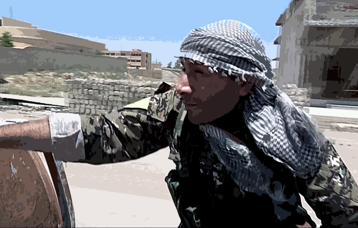 La imagen muestra a un hombre vestido con un uniforme militar y una kefía (una especie de pañuelo) en la cabeza. Él parece estar en un entorno urbano, posiblemente en un área de conflicto. El fondo incluye edificios y una zona que parece abandonada o dañada. El hombre tiene una expresión concentrada y parece estar en alerta, posiblemente observando o preparándose para algo en el entorno que lo rodea.
