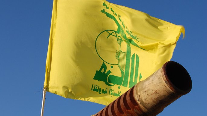 L'image montre un canon pointant vers le ciel, avec un drapeau jaune flottant au-dessus. Sur le drapeau, il y a des inscriptions en arabe ainsi qu'un logo symbolique. Le fond est bleu, suggérant une journée ensoleillée. Le canon présente des signes d'usure, avec une surface rouillée.