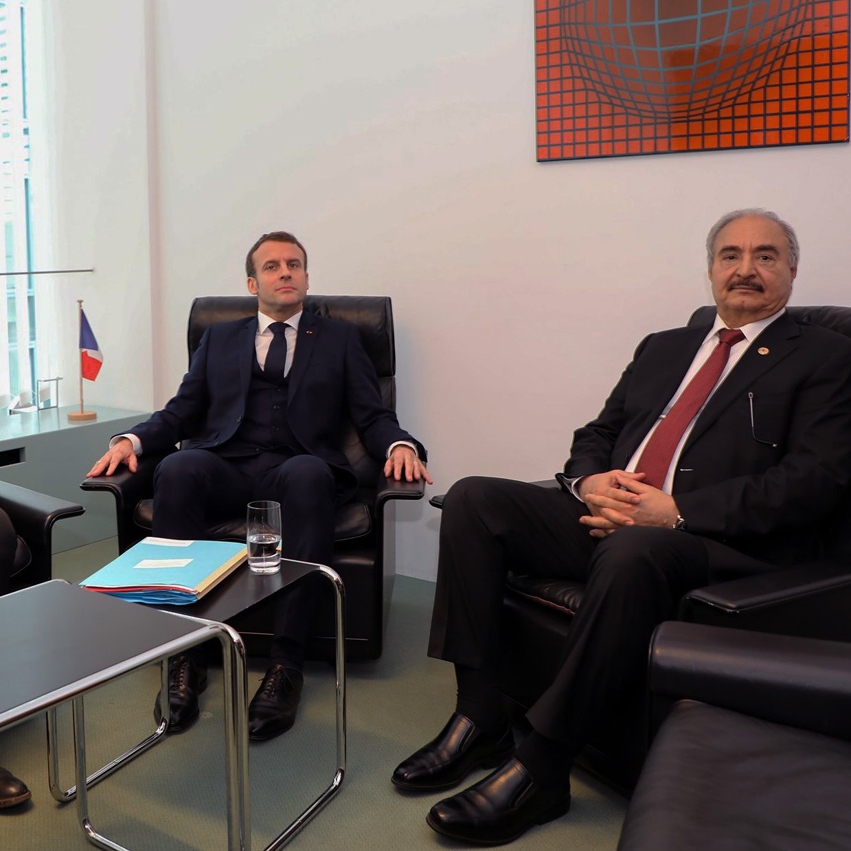 L'image montre trois hommes assis dans un cadre formel. À gauche, un homme avec des cheveux courts, habillé en costume sombre, semble tenir un document. Au centre, un homme en costume bleu regardant droit devant lui. À droite, un homme en costume noir, avec des cheveux grisonnants. Ils sont assis dans des fauteuils confortables, et il y a une table basse entre eux avec un verre d'eau. En arrière-plan, on aperçoit une œuvre d'art et un drapeau français. L'atmosphère semble sérieuse et professionnelle.