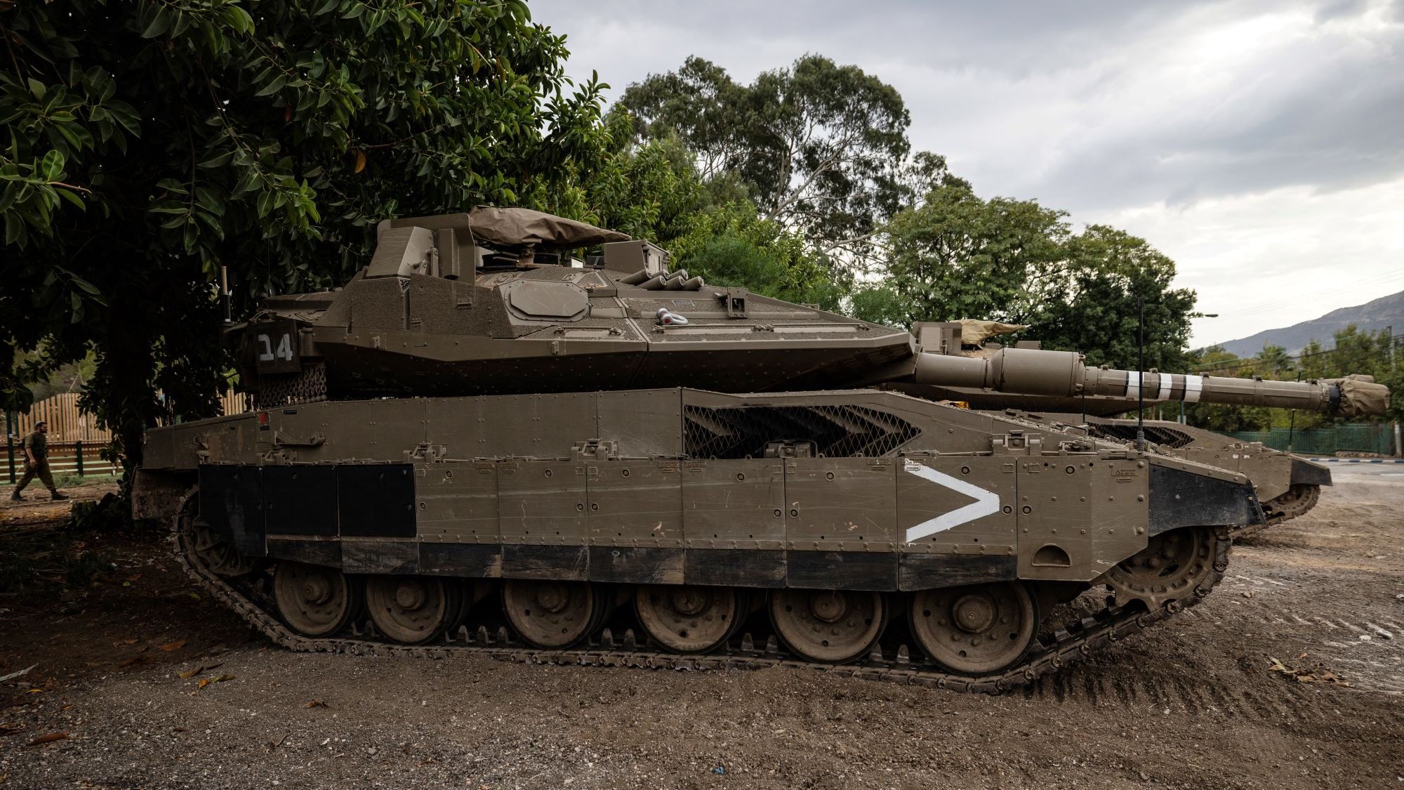 L'image montre un char d'assaut stationné sur un terrain. Le véhicule est de couleur verte foncée et possède des marques de camouflages. Il est équipé d'une tourelle avec un canon pointé vers l'avant. En arrière-plan, on peut voir des arbres et un ciel nuageux, ce qui donne une ambiance militaire à la scène. Le char semble robuste et bien conservé, prêt pour une utilisation éventuelle.