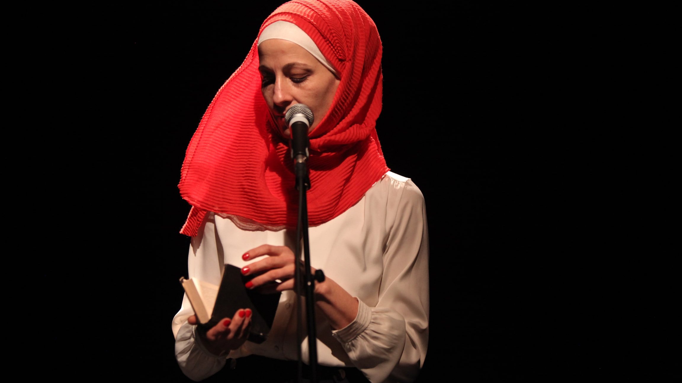 L'image montre une femme debout devant un microphone sur scène. Elle porte un hijab rouge vif et une blouse blanche. Elle tient un petit livre dans ses mains, semblant s'apprêter à lire ou à partager un texte. L'éclairage est sombre, mettant en valeur son visage et l'objet qu'elle tient. L'atmosphère semble intime et concentrée.