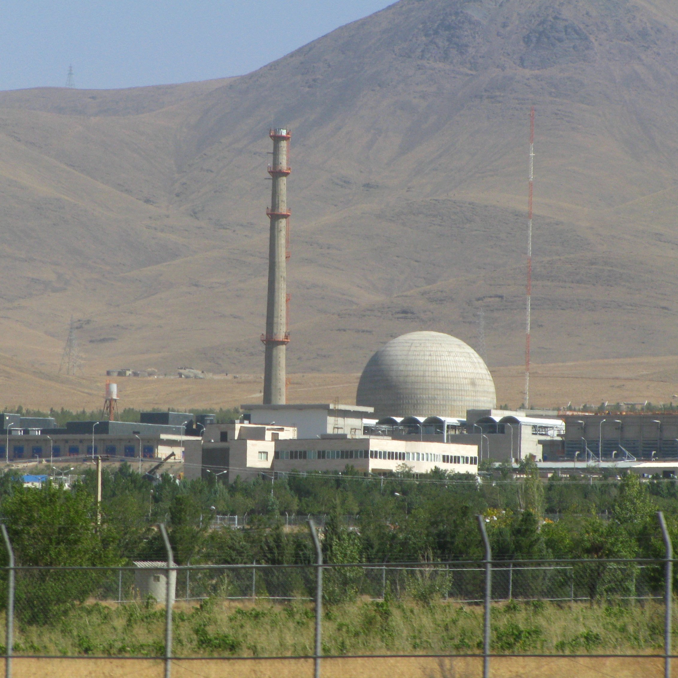 L'image montre une installation industrielle située dans un paysage montagneux. Au premier plan, on peut voir une clôture entourant le site. À l'arrière, il y a un bâtiment principal avec une grande structure en forme de dôme et une cheminée élevée, qui semble émettre de la vapeur ou des gaz. Des antennes et d'autres équipements sont également visibles, tandis que le terrain environnant est semi-aride avec une végétation limitée. Le ciel est dégagé et montagneux, ajoutant à l'aspect éloigné de l'installation.