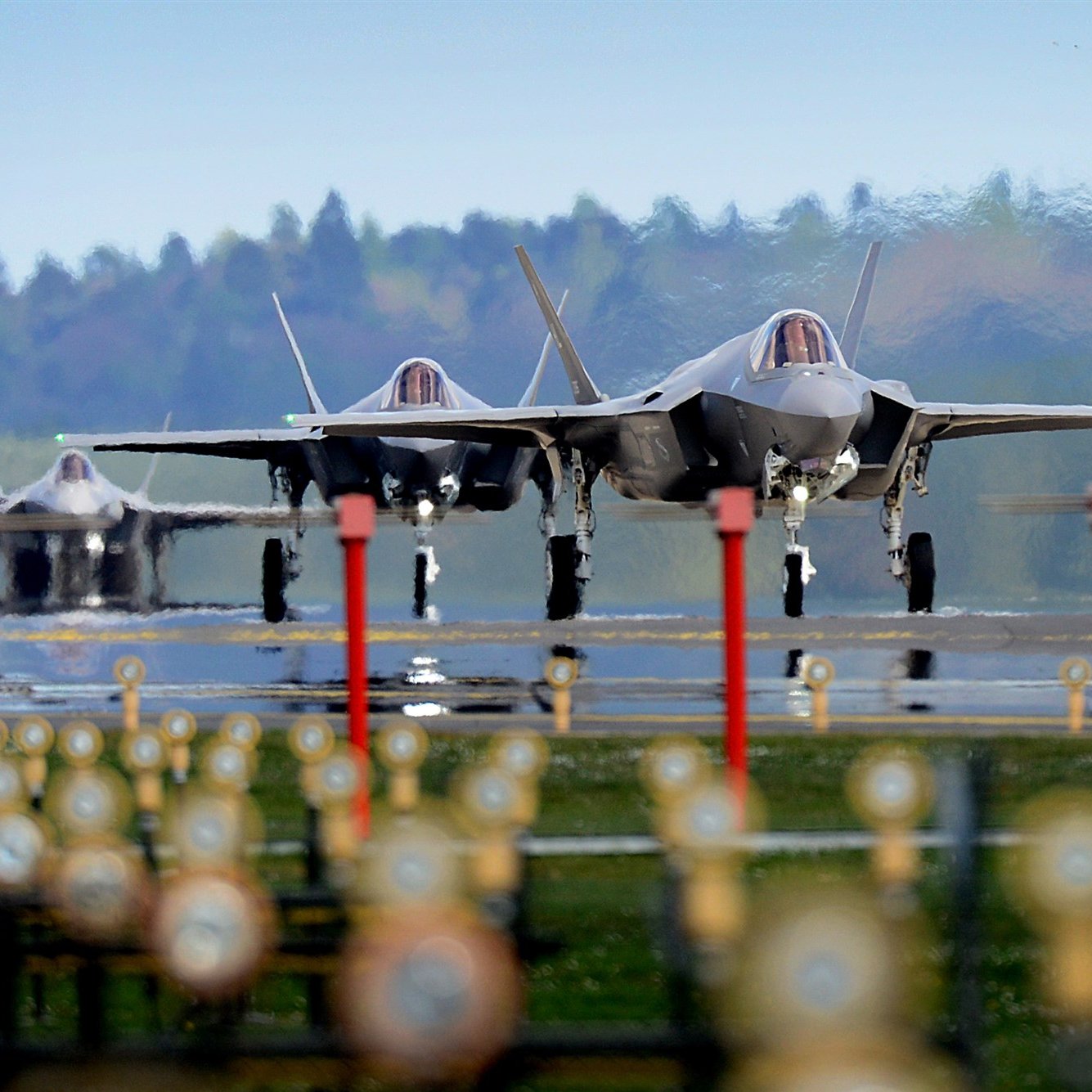 L'image montre plusieurs avions de chasse F-35 alignés sur une piste d'atterrissage. Au premier plan, on peut voir des lumières et des repères au sol, tandis que les avions sont prêts au décollage. En arrière-plan, on aperçoit une forêt verdoyante sous un ciel clair. L'ambiance général semble être celle d'une base aérienne, mettant en avant la puissance et la technologie militaire moderne.