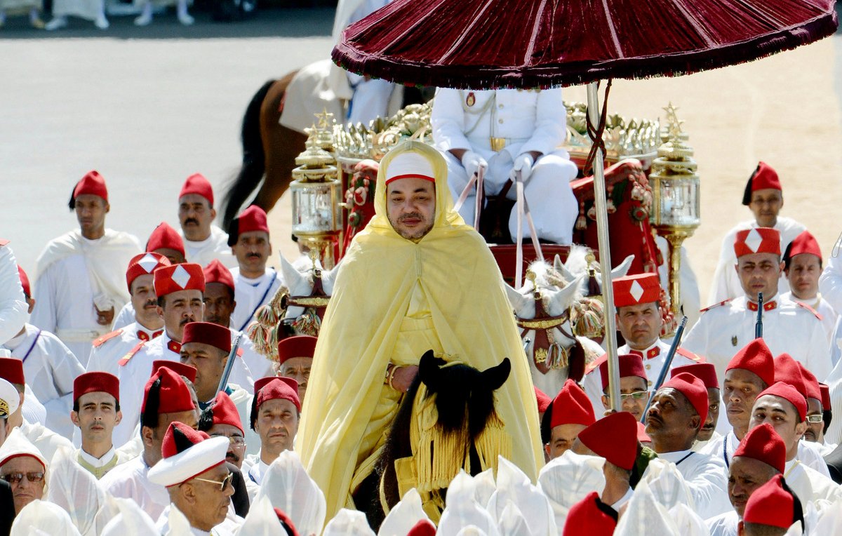 L'image montre un roi entouré de nombreux participants, tous vêtus de vêtements traditionnels. Le roi est monté sur un cheval, revêtu d'une cape jaune et portant un chapeau distinctive. Au-dessus de lui se trouve un parasol rouge. Les membres de l'entourage portent des turbans rouges, tandis que d'autres sont habillés en blanc. L'arrière-plan semble être une cérémonie officielle, avec des spectateurs et un décor sobre. Cette scène évoque une ambiance de tradition et de solennité.