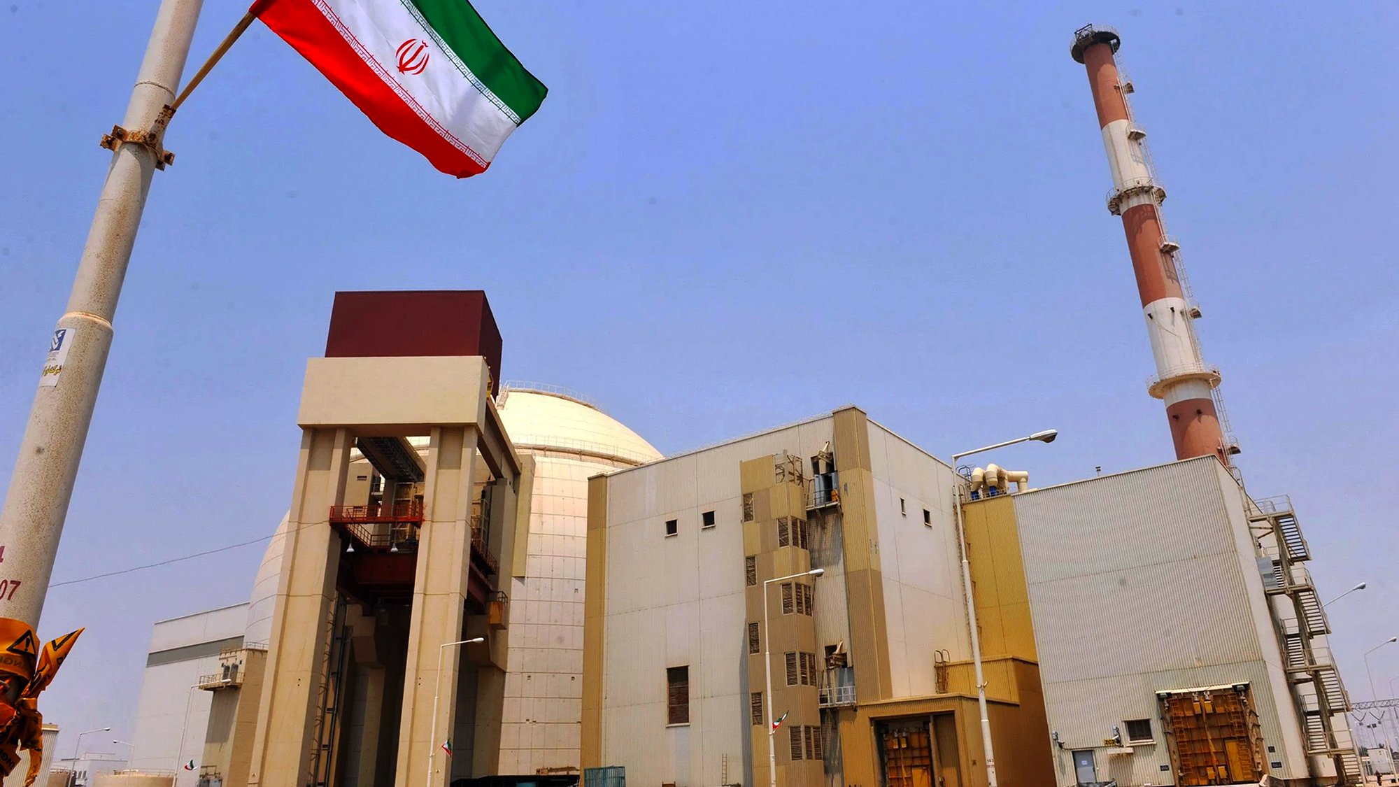 در این تصویر، یک تأسیسات هسته‌ای در ایران دیده می‌شود. در سمت چپ تصویر، پرچم ایران در حال برافراشته شدن است. ساختمان‌ها و برج‌های دودکش در پس‌زمینه قرار دارند و آسمان آبی و شفاف است. تأسیسات به نظر صنعتی و مدرن می‌آیند و نشان‌دهنده فعالیت‌های مرتبط با انرژی هسته‌ای هستند.