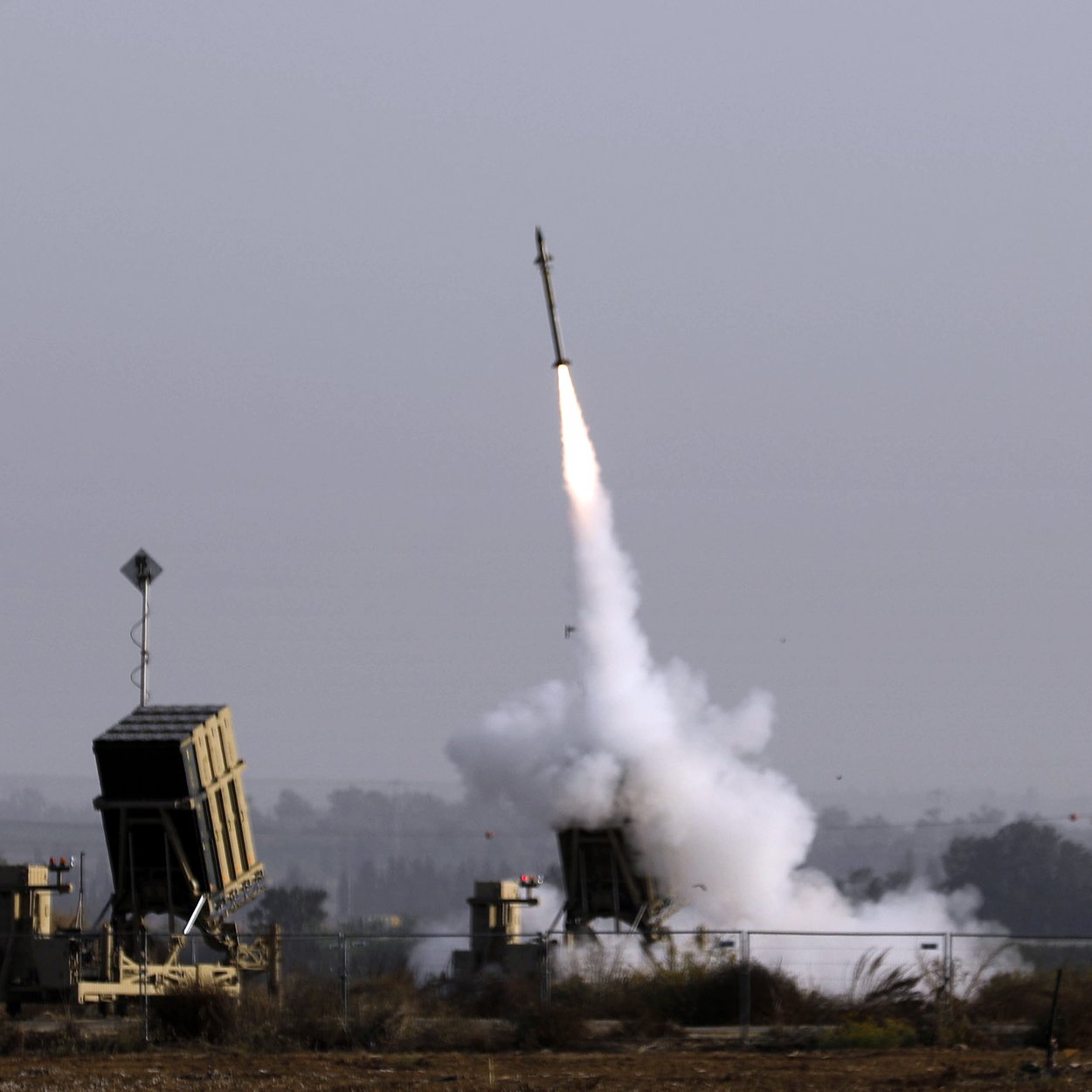 L'image montre un système de défense antimissile en action, avec un missile tiré vers le ciel. On peut voir les lanceurs de missiles, qui sont situés sur un terrain ouvert, entourés de végétation. Une traînée de fumée s'élève du lanceur, indiquant le moment du lancement. Le ciel est clair, avec une légère brume qui lui confère une atmosphère chargée.