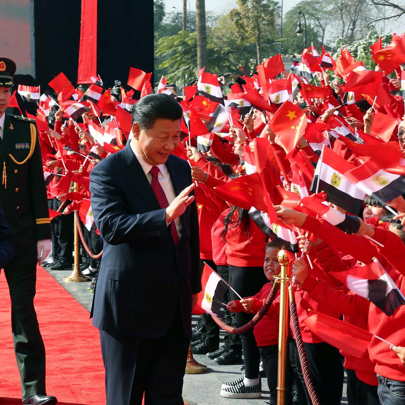 در این تصویر، یک مراسم رسمی در حال برگزاری است. فردی با کت و شلوار تیره و عینک آفتابی در سمت چپ تصویر قرار دارد و به نظر می‌رسد که یک مقام دولتی است. در سمت راست، مرد دیگری که لباس رسمی پوشیده، با لبخند به سمت جمعیتی که پرچم‌های چین و مصر را در دست دارند، نزدیک می‌شود. جمعیت در حال موج زدن به پرچم‌ها و ابراز شادی هستند. زمین پوشیده از فرش قرمز است و تعدادی از نیروهای نظامی نیز در پس‌زمینه دیده می‌شوند. فضای کلی مراسم بسیار رسمی و پرانرژی به نظر می‌رسد.