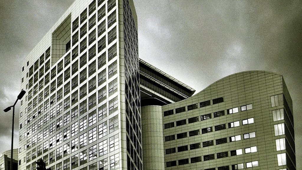 L'image montre un bâtiment moderne avec une architecture contemporaine. La structure est composée de plusieurs angles et surfaces réfléchissantes, donnant une impression de sophistication. Les vitres sont grandes, et le design présente des lignes épurées. Le ciel en arrière-plan est nuageux, ce qui peut créer une ambiance dramatique. Les tons de l'image sont en noir et blanc, accentuant les contrastes entre la lumière et l'ombre sur la façade du bâtiment.