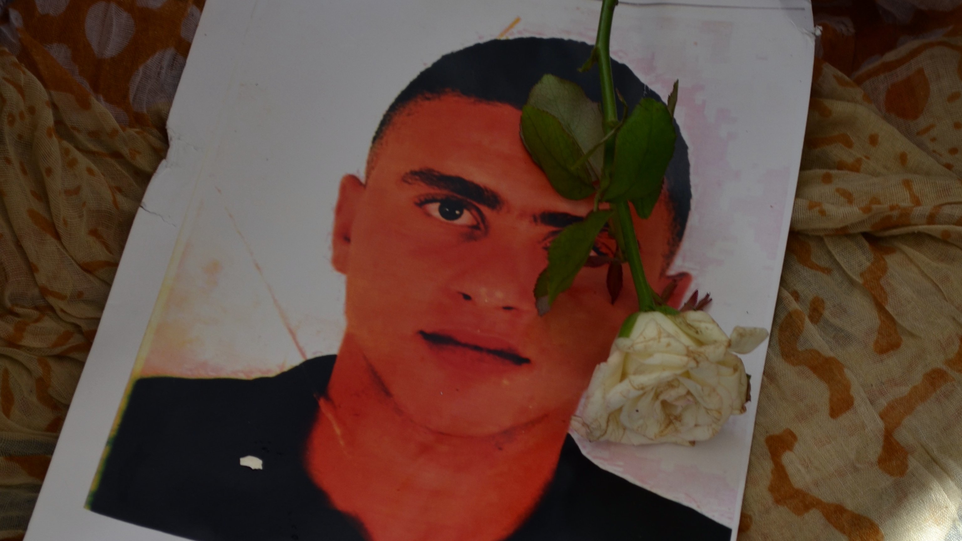 L'immagine mostra un ritratto di un giovane uomo posato su un tessuto colorato. Sopra il ritratto è adagiata una rosa bianca, che aggiunge un tocco di dolcezza e commemorazione. Il contrasto tra il volto del ragazzo e il fondo morbido e multicolore crea un'atmosfera di riflessione e rispetto.