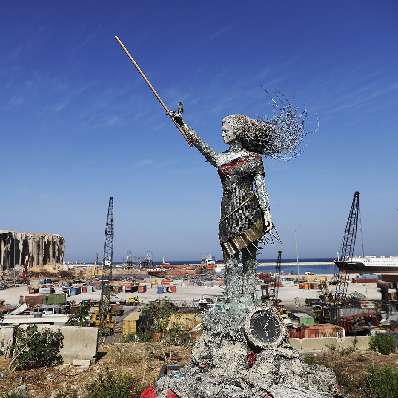 L'image présente une sculpture en métal d'une femme levant un bâton, symbolisant probablement la force et la résilience. Elle se tient sur une plateforme, surplombant un paysage industriel ou portuaire, avec des conteneurs, des grues et des bâtiments en arrière-plan. On peut également apercevoir des vestiges d'un bâtiment détruit, suggérant une scène de reconstruction ou de renouveau après une catastrophe. Le ciel est dégagé et ensoleillé, contrastant avec l'environnement chaotique du port.