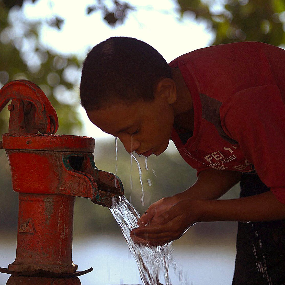 Sur l'image, un jeune garçon penche sa tête en avant pour recueillir de l'eau qui jaillit d'une pompe manuelle rouge. Il a l'air concentré et utilise ses mains pour attraper l'eau qui s'écoule. En arrière-plan, on peut apercevoir un paysage flou avec un plan d'eau. La lumière semble douce, et l'atmosphère évoque un moment de fraîcheur et de vie quotidienne.