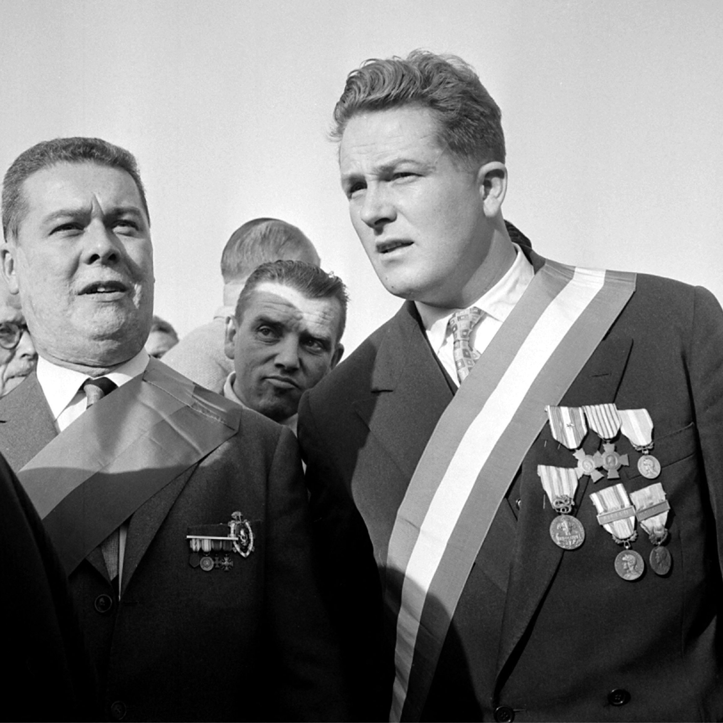 L'image montre deux hommes qui semblent participer à un événement officiel ou une cérémonie. L'un des hommes porte plusieurs médailles accrochées à sa veste, ainsi qu'une écharpe. L'autre homme porte également une écharpe, mais il a moins de médailles visibles. L'ambiance paraît solennelle, avec d'autres personnes en arrière-plan. Les expressions des deux hommes suggèrent qu'ils sont engagés dans une conversation ou qu'ils se concentrent sur l'événement.