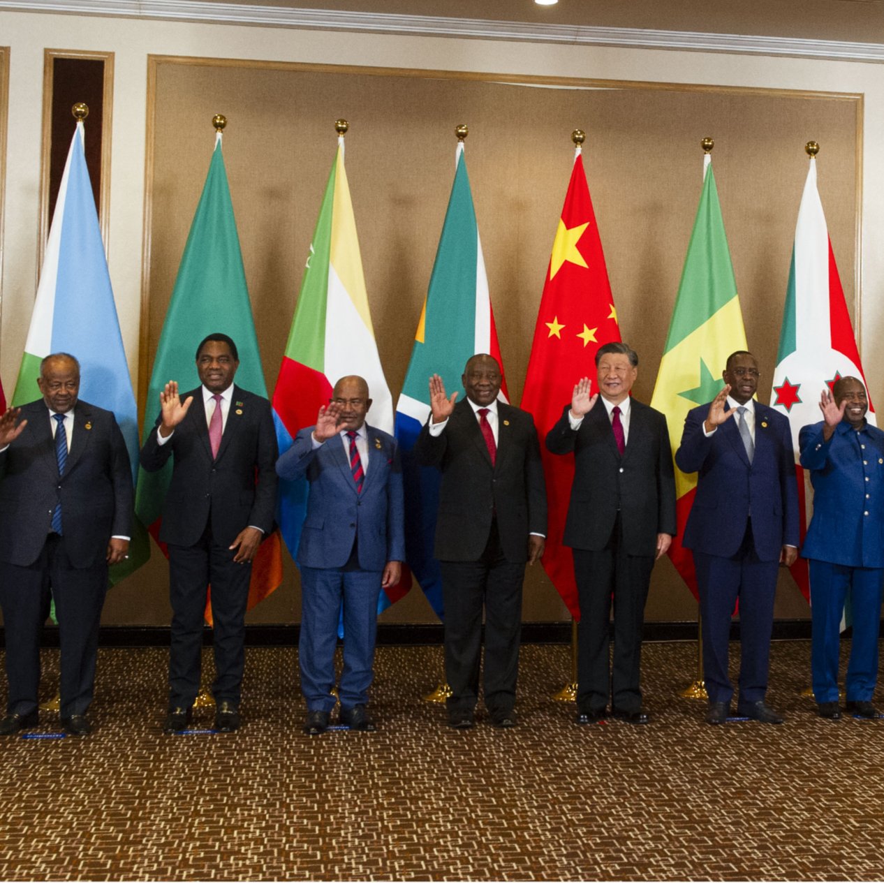 تظهر الصورة مجموعة من القادة أو الممثلين الرسميين من دول مختلفة يقفون في صف واحد. خلفهم، تُعلق أعلام تمثل الدول أو المنظمات المختلفة، بما في ذلك علم الاتحاد الأفريقي. يبدو أن الجميع يلوح بيديه في تعبير عن التحية. يرتدي القادة بدلات رسمية، وبعضهم يرتدي الملابس التقليدية. تعكس الصورة مناسبة رسمية أو قمة تجمع بين الدول.