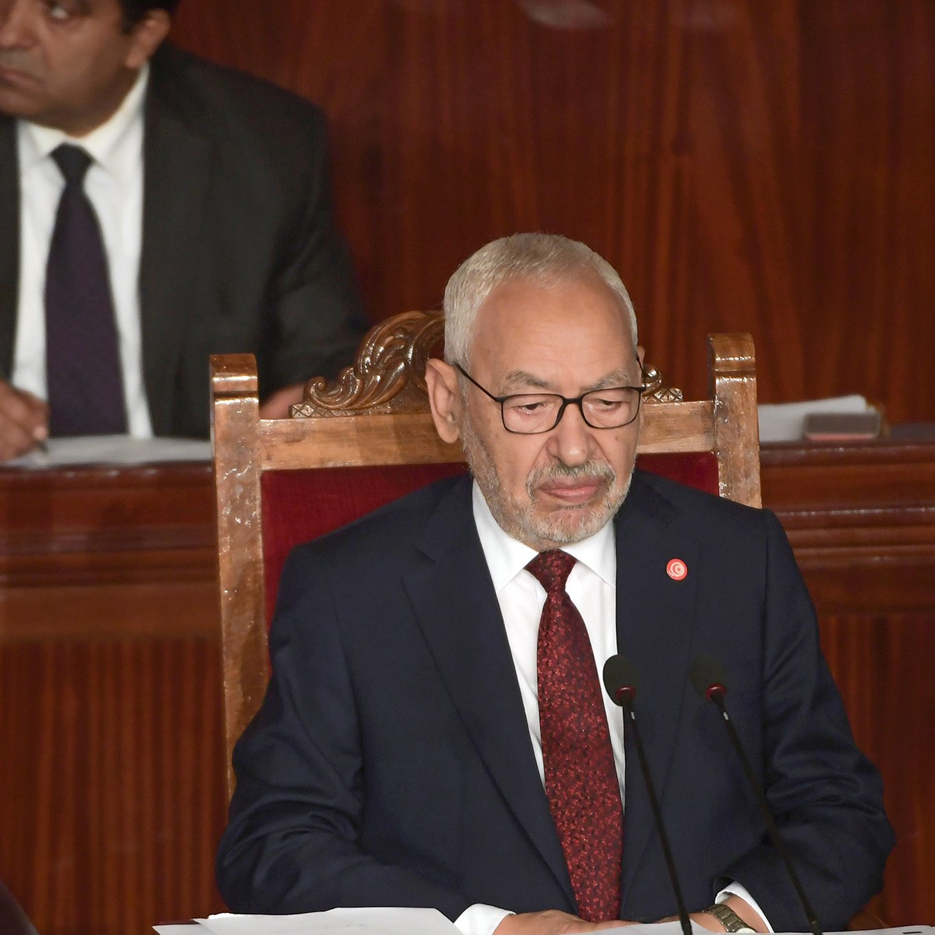 L'image montre un homme assis à un bureau, portant un costume sombre et des lunettes. Il semble être en train de s'exprimer ou de lire un document, avec une expression sérieuse. À l'arrière-plan, une autre personne est visible, vêtue d'un costume, observant la scène. La pièce a des murs en bois et semble être un cadre formel, probablement une assemblée ou un parlement.