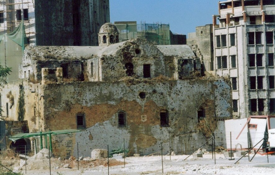L'image montre des ruines d'un bâtiment ancien, probablement une église, dont la structure est en pierre et partiellement en décomposition. La façade est endommagée et recouverte de végétation, tandis qu'en arrière-plan, on peut voir des immeubles modernes en construction ou en rénovation. Le contraste entre l'ancien et le moderne donne une atmosphère de dégradation et de changement architectural. La zone semble être en chantier, avec des barrières et des échafaudages visibles.