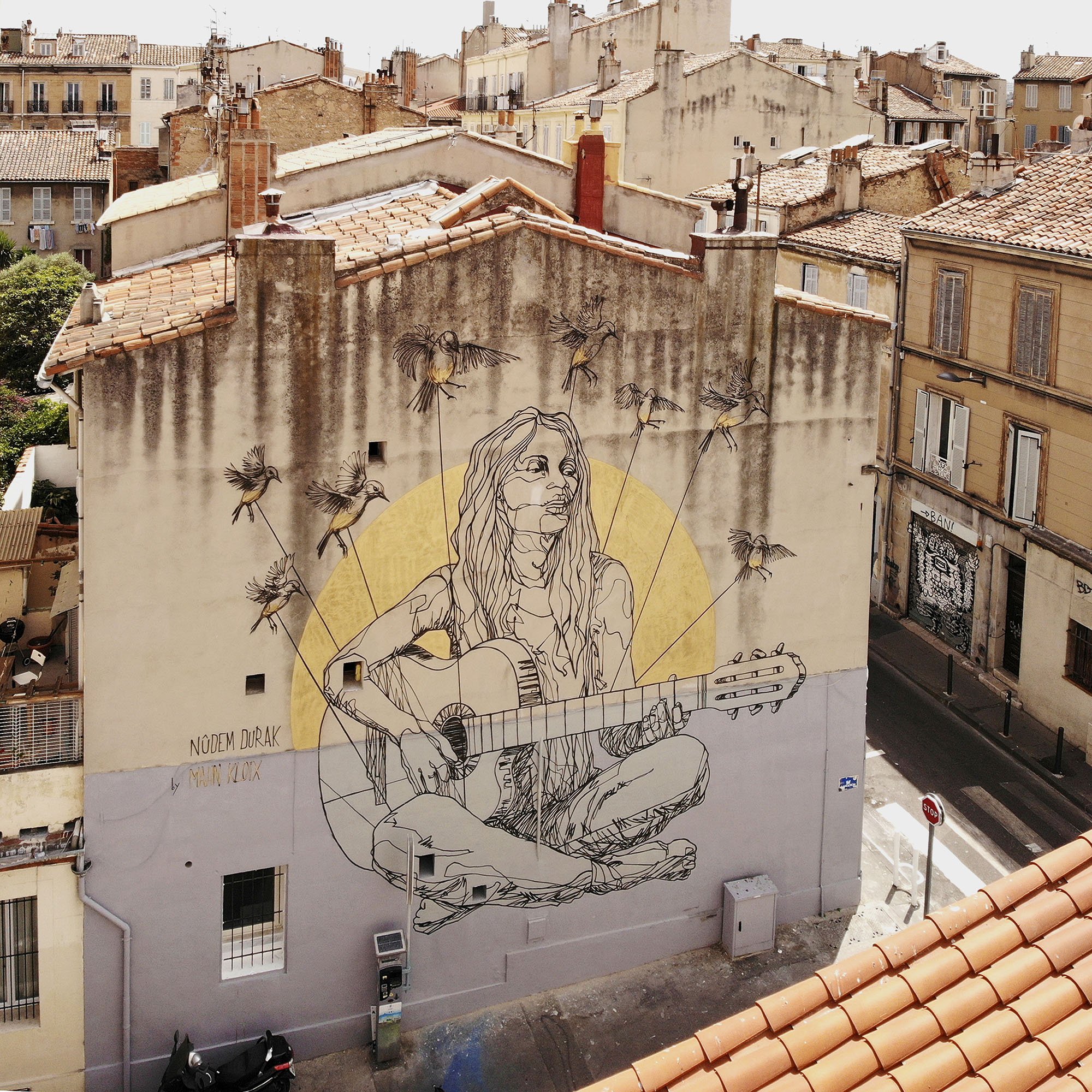 L'image montre une fresque murale colorée sur un bâtiment. Au centre, on voit une représentation d'une jeune femme assise, jouant de la guitare. Elle a de longs cheveux et un look décontracté. Autour d'elle, il y a plusieurs abeilles illustrées. En arrière-plan, on aperçoit des bâtiments typiques avec des fenêtres et des toits d'une ville. La fresque semble apporter une ambiance artistique et vivante à l'environnement urbain.
