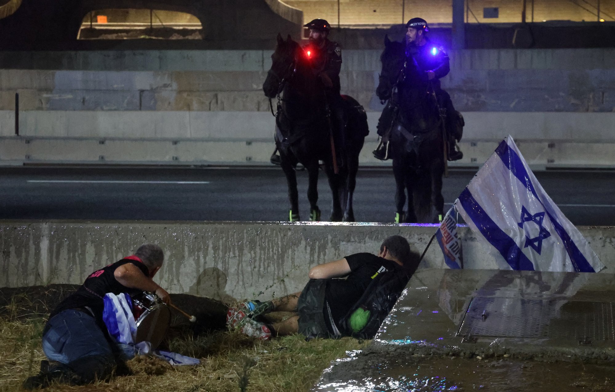 تظهر الصورة مشهدًا في مدينة ما، حيث يتواجد رجلان على الأرض بجانب ممر مائي أو خندق. أحدهما يبدو أنه على الأرض بينما الآخر يختبئ خلف حواجز. في الخلفية، يظهر اثنان من رجال الشرطة على ظهور الخيل، مجهزين بأضواء ملونة على خوذتهم. هناك أيضًا علم إسرائيلي ملقى على الأرض بالقرب من الرجلين. الجو العام يوحي بوجود حدث أو مواجهة تتعلق بالتوترات الاجتماعية أو السياسية.