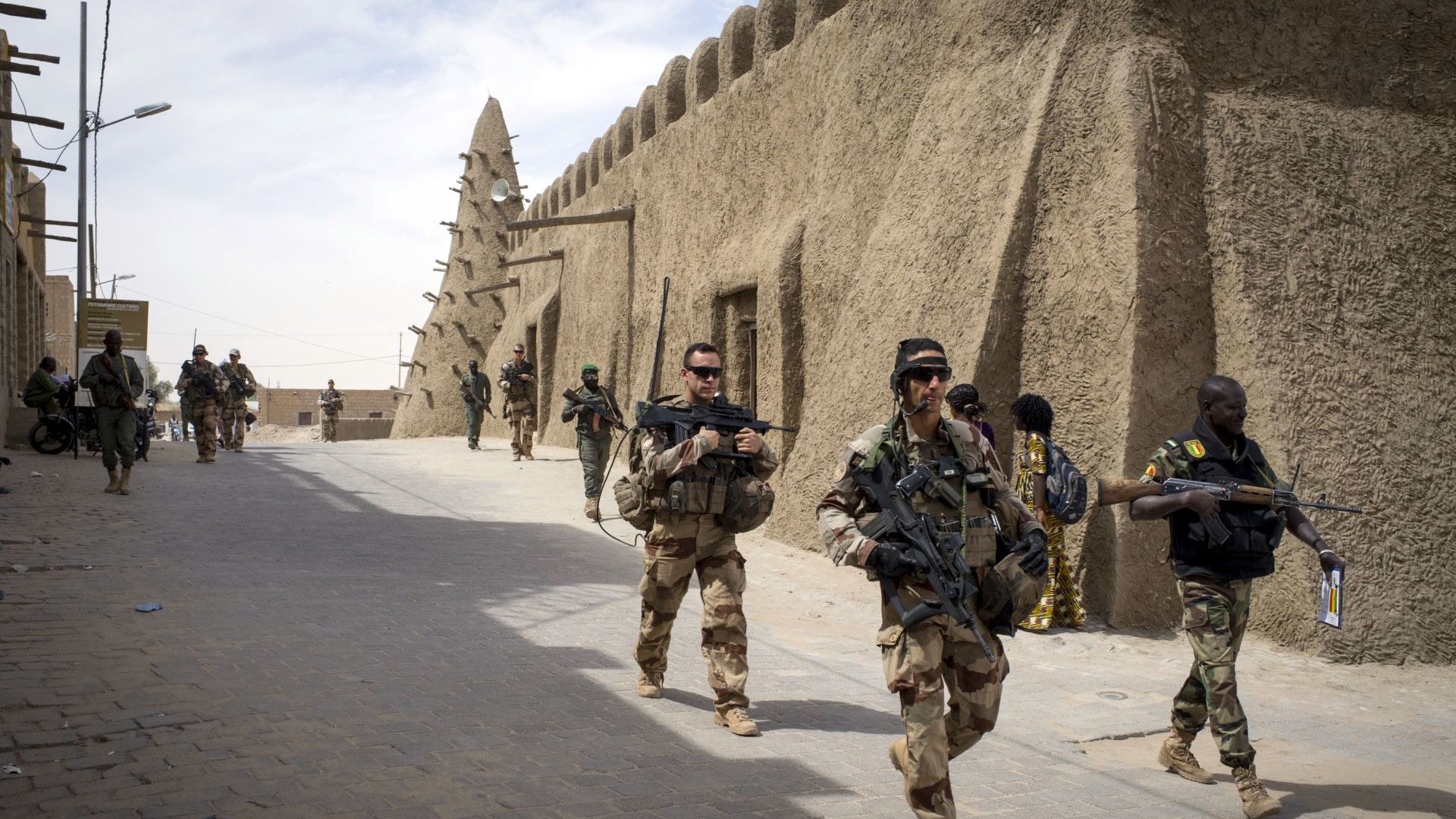 تظهر الصورة جنوداً يسيرون في شوارع منطقة تاريخية. المباني في الخلفية تبدو مصنوعة من الطين، وتمتاز بتصميم تقليدي. الجنود يحملون أسلحة ويبدو عليهم التركيز والحذر. يتواجد أيضاً شخصيات أخرى في الخلفية، مما يدل على وجود نشاطات متعددة في المنطقة. السماء تبدو صافية مع بعض السحب.