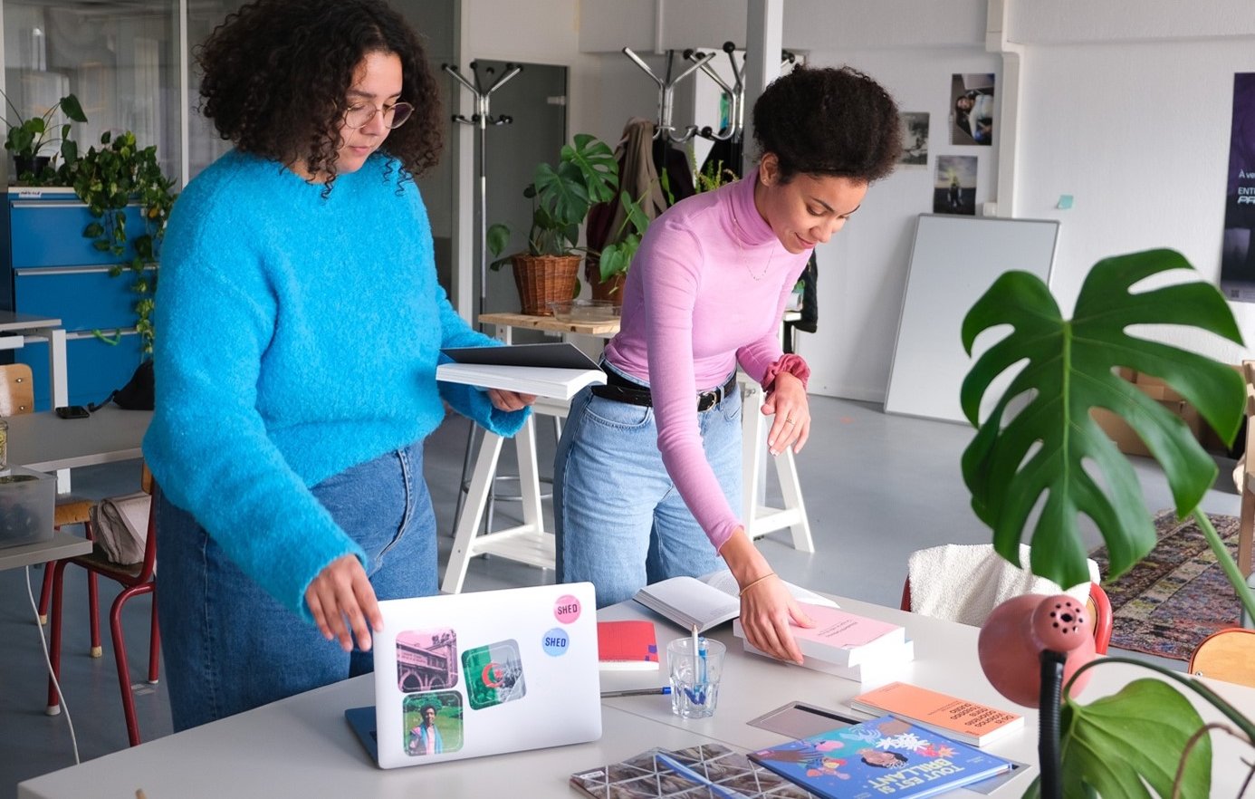 The image shows two women in a modern workspace, engaged in an activity together. One woman, wearing a bright blue sweater, is standing and holding a stack of papers or a folder. The other woman, dressed in a pink top, is bending over a table, arranging or placing items. The table is cluttered with books, a laptop, and other materials, indicating a creative environment. There are plants in the background, adding a touch of greenery to the space. The overall atmosphere appears collaborative and productive.