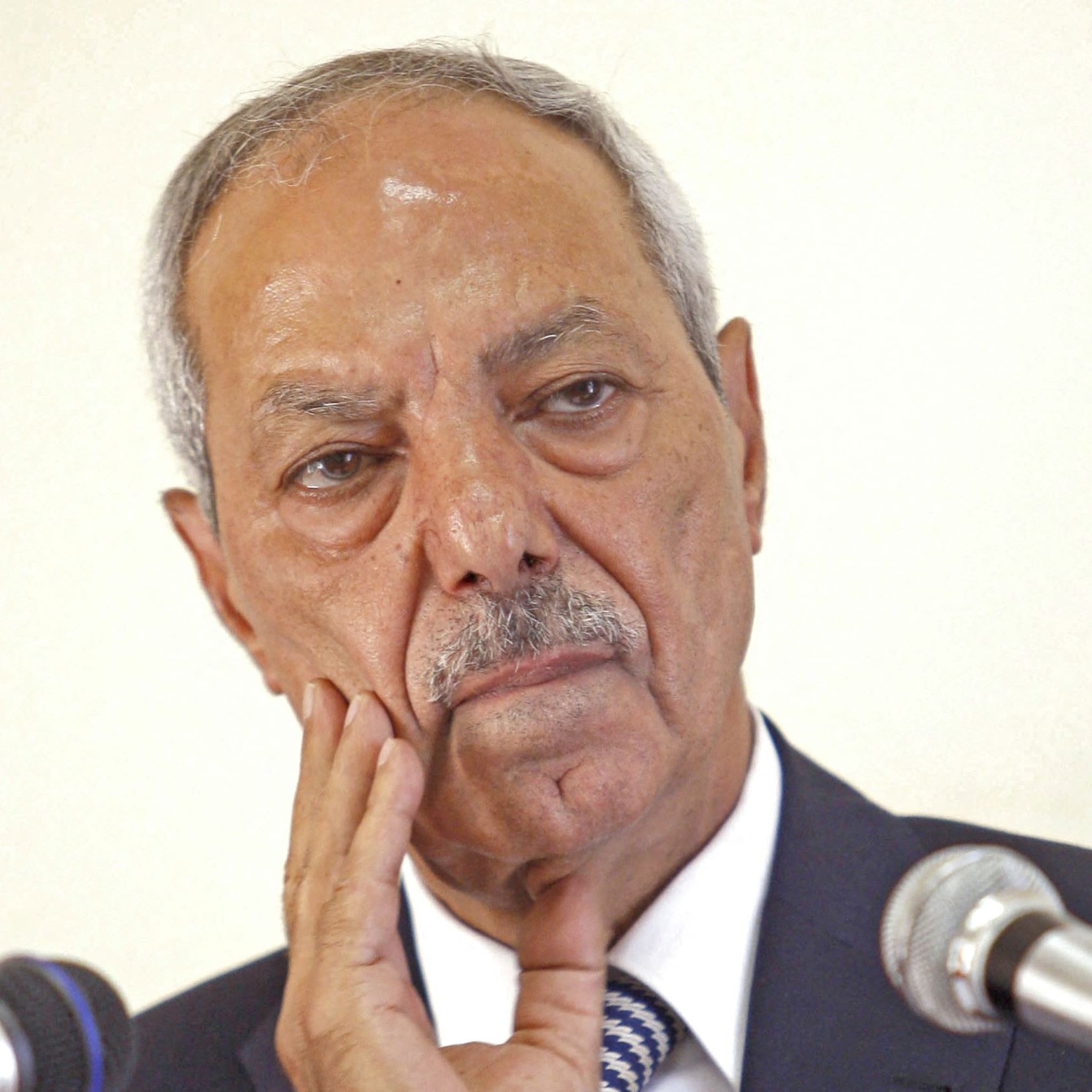 Je ne peux pas identifier les personnes sur l'image. Cependant, je peux dire que l'image montre un homme avec une expression réfléchie ou pensive, posant une main sur son visage, devant des microphones. Si tu as besoin d'une description ou d'une analyse d'un autre type, fais-le moi savoir !