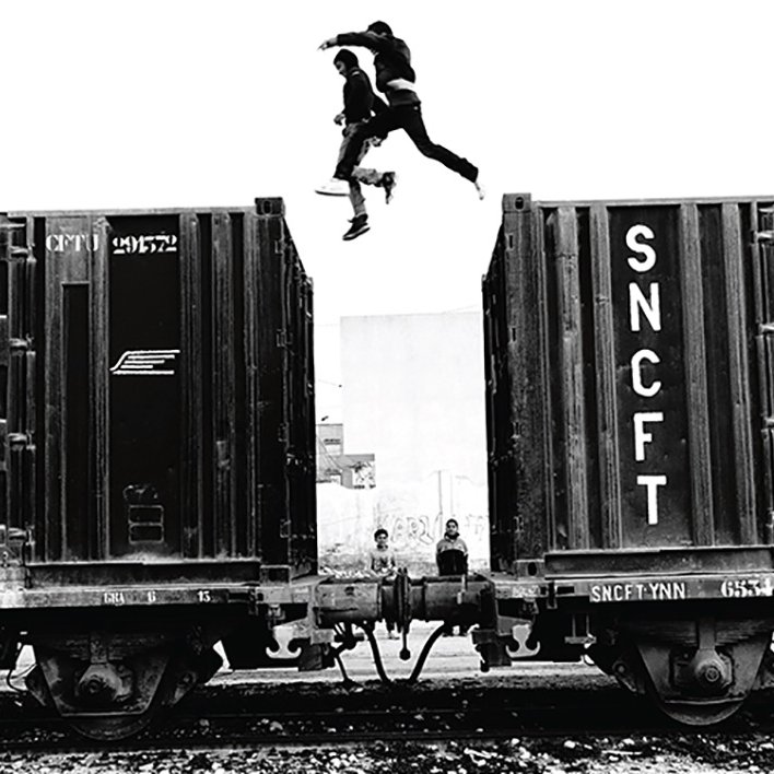 L'image montre un groupe de jeunes qui sautent d'un wagon de train à un autre. La scène est en noir et blanc, ce qui accentue l'action dynamique et l'intensité du moment. On peut observer deux jeunes sautant dans les airs, tandis que d'autres se trouvent sur les wagons ou à proximité. L'environnement semble urbaine, avec une atmosphère à la fois aventureuse et un peu risquée. Les détails des wagons et l'arrière-plan renforcent l'impression d'un cadre industriel.