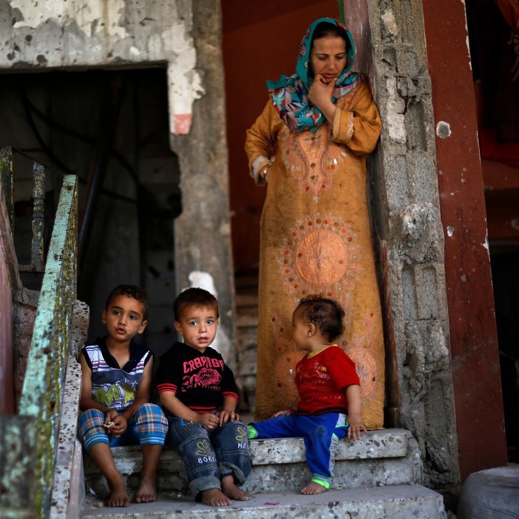 L'image montre une femme et plusieurs jeunes enfants assis sur des marches en béton. La femme est vêtue d'une robe longue, tandis que les enfants, assis près d'elle, portent des vêtements colorés. Le décor environnant semble ancien et en mauvais état, avec des murs écaillés et des escaliers. L'ambiance générale évoque une atmosphère de vie quotidienne dans un environnement modeste.