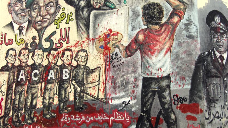 L'image montre un mural coloré, rempli de graffitis et d'illustrations. On y voit une scène dynamique où une personne, de dos, est en train de peindre avec un pinceau, probablement en train d'exprimer un message de frustration ou de résistance. Autour de cette figure, il y a diverses représentations de policiers et de figures autoritaires, certains avec des visages mécontents, indiquant une atmosphère de contestation. Le mur est éclaboussé de peinture rouge, ce qui pourrait symboliser la violence ou le sang, renforçant le message de lutte contre la répression. Les lettres "ACAB" sont visibles, ce qui fait référence à une phrase souvent associée à la critique des forces de police. L'ensemble de la composition semble porter un message fort sur l'opposition à l'autorité.