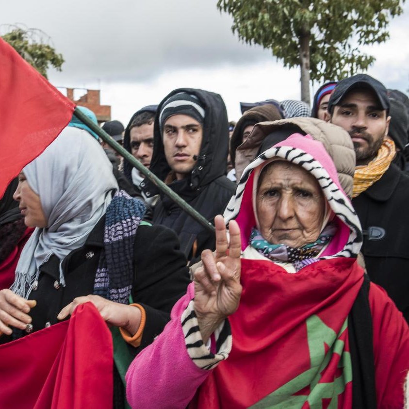 The image depicts a group of people participating in a demonstration. In the foreground, an older woman holds a flag featuring the Moroccan national emblem and makes a peace sign with her hand. She is surrounded by others who are also holding flags and banners, all dressed for colder weather. The atmosphere suggests a sense of solidarity among the demonstrators, with the crowd positioned against a backdrop of cloudy skies.