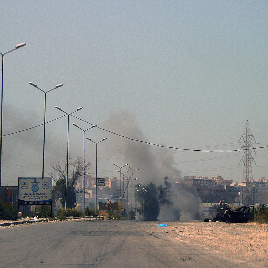 L'image montre une scène de route apparemment désertée, avec quelques bâtiments sur les côtés. On peut apercevoir de la fumée s'élevant au loin, indiquant probablement un incendie ou une explosion. Des panneaux de signalisation sont visibles, ainsi que des lampadaires le long de la route. L'atmosphère semble chargée et la visibilité est atténuée par la fumée.