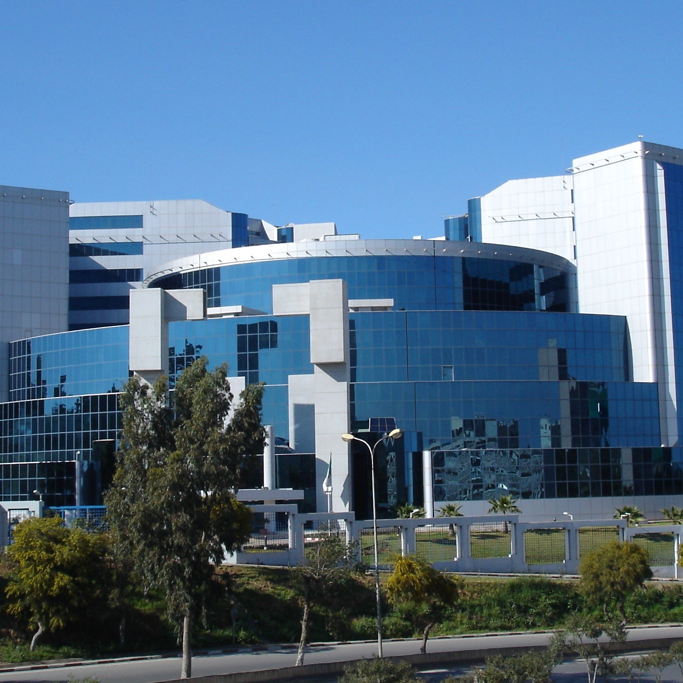 الصورة تظهر مبنى حديث التصميم، يتكون من واجهة زجاجية تعكس الضوء، مما يعطيه مظهراً عصريًا. يحتوي المبنى على عدة طوابق وزوايا مختلفة تجعله بارزًا في المشهد. تحيط به أشجار ونباتات، مما يضيف لمسة من الطبيعة إلى التصميم المعماري. السماء زرقاء وصافية، مما يعزز جمال الصورة.