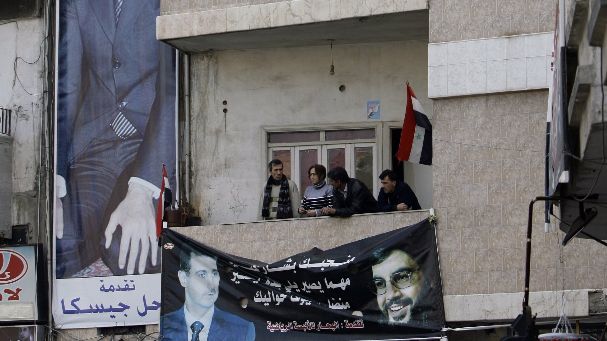L'image montre un bâtiment avec une grande affiche représentant un homme en costume, probablement une figure politique. On peut voir plusieurs personnes sur un balcon au premier étage, observant l'extérieur. En dessous, il y a une bannière affichant un message, et des drapeaux sont visibles, ce qui suggère un contexte politique ou de soutien à une cause particulière. Les murs du bâtiment sont décorés de manière simple, et l'ambiance semble être formelle.