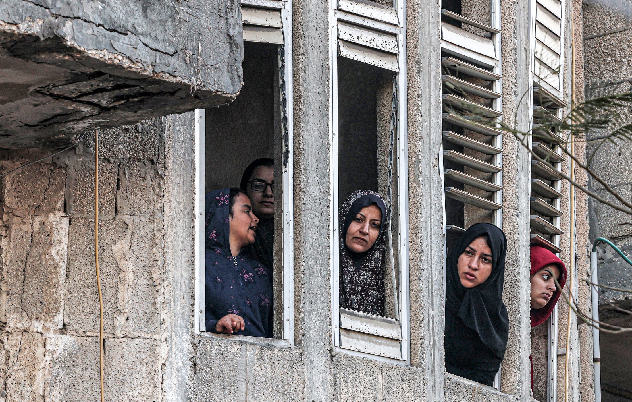 The image shows several women looking out of narrow windows in what appears to be a multi-story concrete building. The women are wearing headscarves and appear to be observing something outside with serious or concerned expressions. The building's exterior is worn and weathered, with visible signs of age and damage. The women are closely peering through the open windows, suggesting that they are trying to get a better view of something important happening outside. The overall mood of the image seems tense or somber.