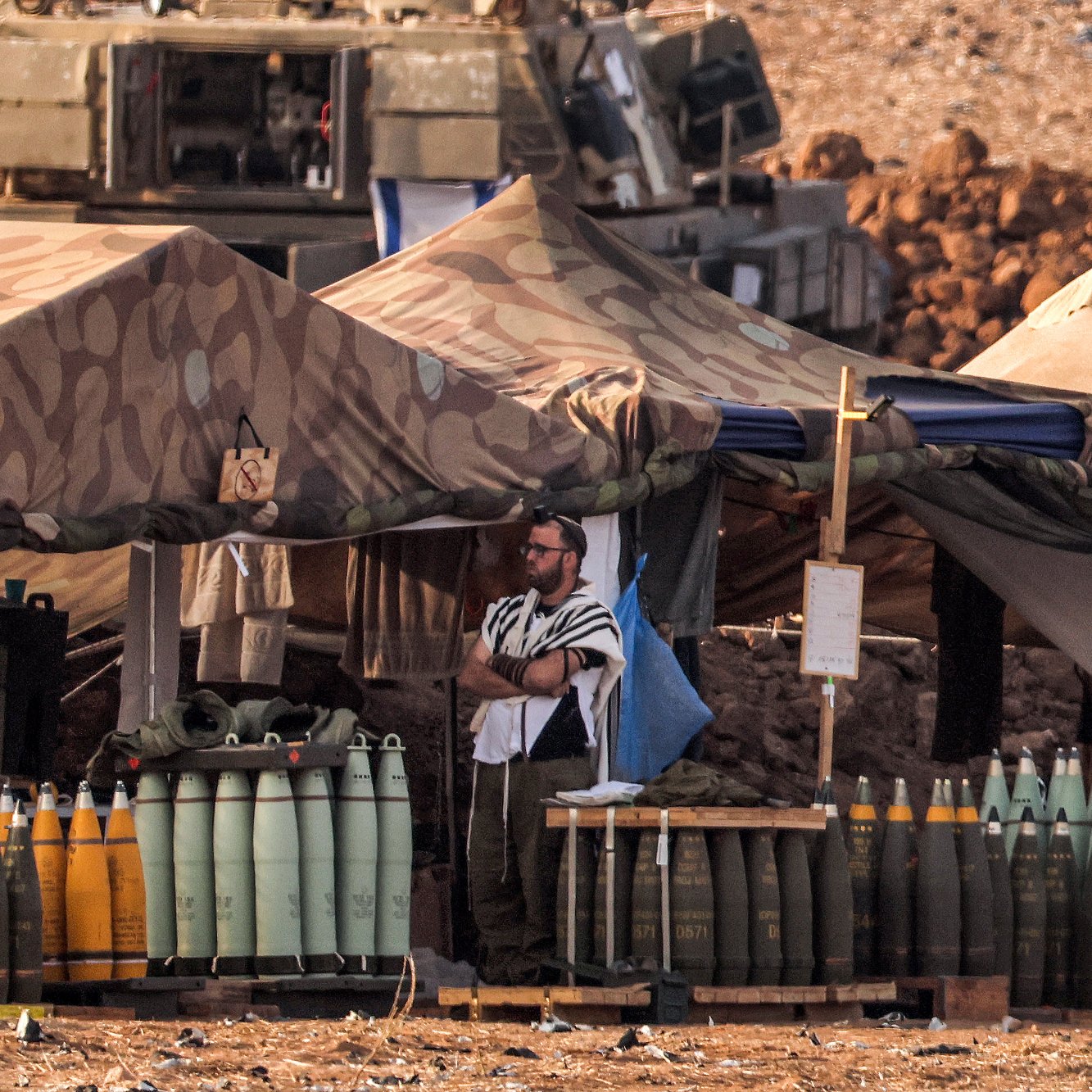 L'image montre un camp militaire en plein air. On y voit des tentes camouflées, et plusieurs obus de différentes tailles sont alignés sur des palettes. Un homme, possiblement en tenue religieuse, se tient debout près des tentes. En arrière-plan, on aperçoit un véhicule militaire. Le sol est poussiéreux, et le paysage semble aride, indiquant une zone de conflit ou d'entraînement militaire.